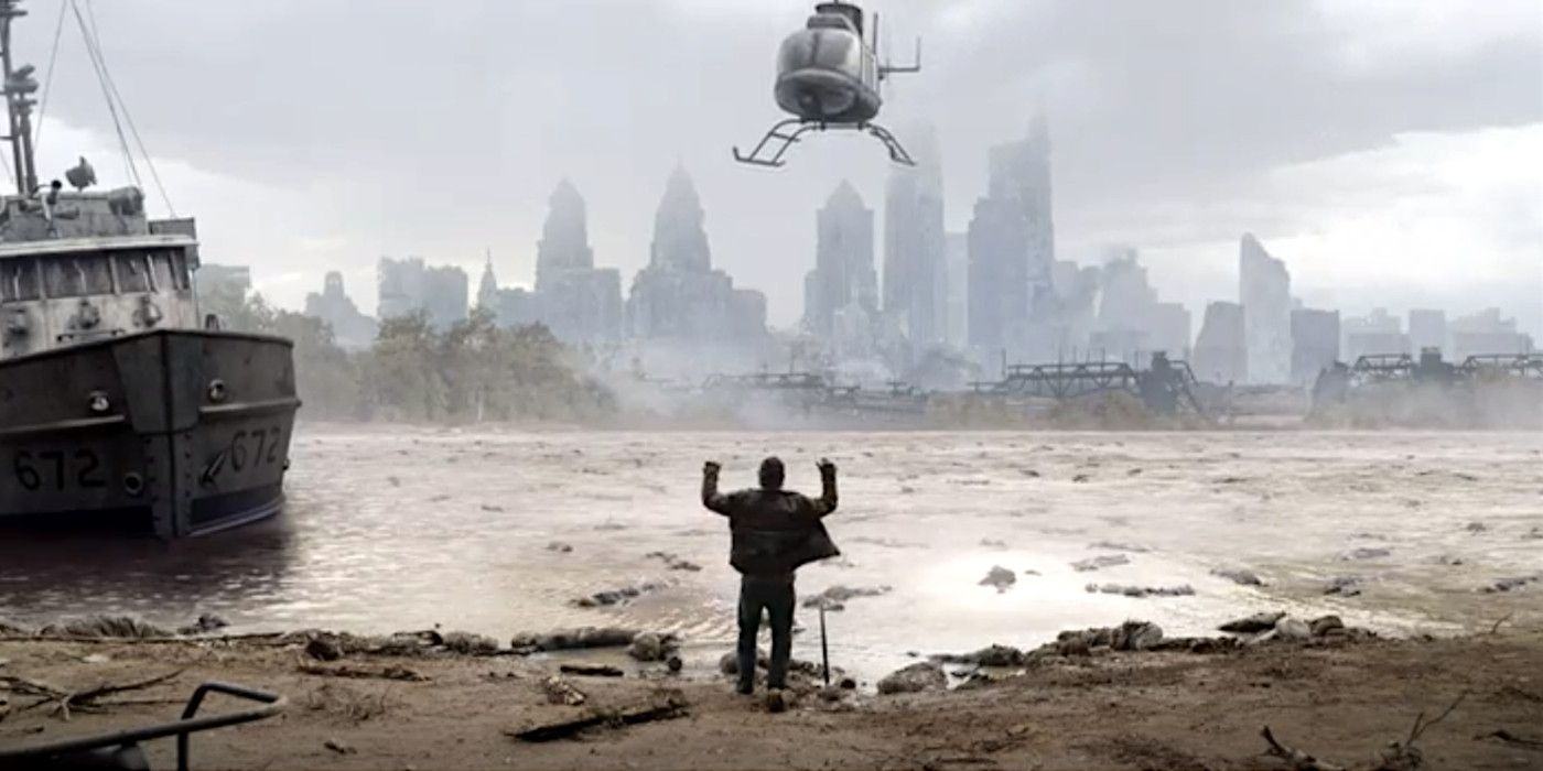 A person stands on a muddy riverbank with his arms raised as a helicopter hovers above him with the blurred city skyline in the distance